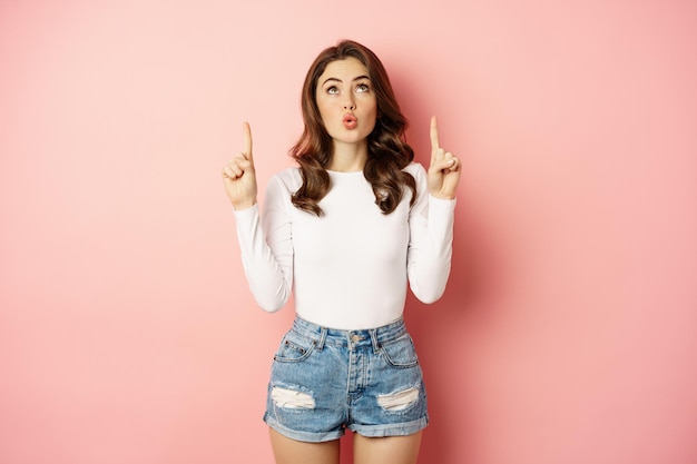 Retrato de mulher caucasiana surpresa, garota estilosa apontando os dedos para cima e parecendo impressionada, mostrando o logotipo da loja, anúncio de desconto, fundo rosa