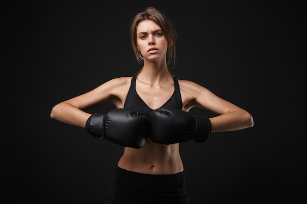 Retrato de mulher caucasiana tensa em roupas esportivas, posando para a câmera com luvas de boxe durante treino no ginásio isolado sobre fundo preto