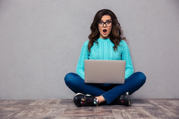 Retrato de mulher chocada, sentada no chão com o laptop e na parede cinza