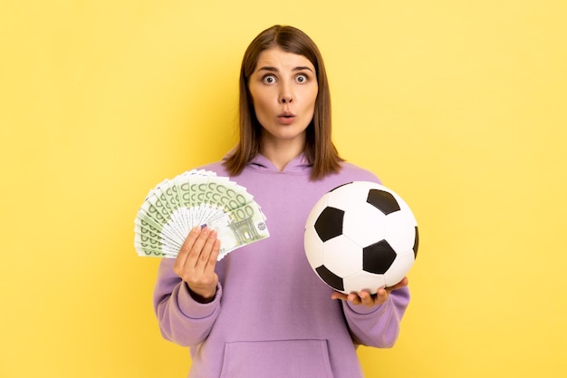 Retrato de mulher com a boca aberta segurando uma bola de futebol e fã de euro espantado com apostas de campeonato de futebol usando capuz roxo Foto de estúdio indoor isolada em fundo amarelo