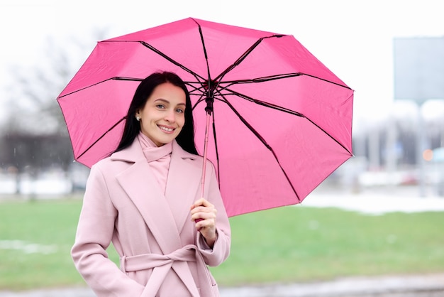 Retrato de mulher com casaco que fica na chuva e guarda-chuva rosa.