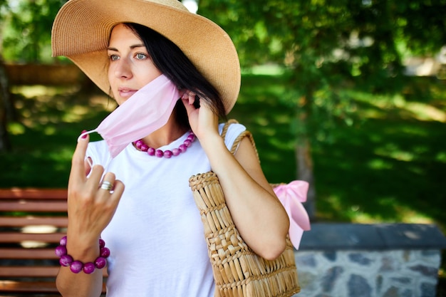 Retrato de mulher com chapéu de palha tira a máscara protetora rosa, no parque ao ar livre na cidade, conceito de autocuidado, vida durante a pandemia de coronavírus, quarentena