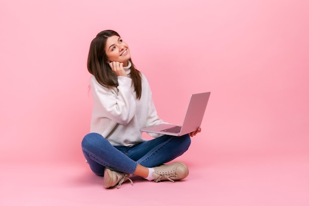 Retrato de mulher com expressão facial sonhadora sentada com as pernas cruzadas no chão segurando o laptop