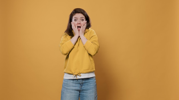 Retrato de mulher com medo fazendo gesto com a mão, tocando o rosto com medo, sentindo medo e medo sobre fundo amarelo. Jovem fazendo careta de rosto chocado sendo assustado no estúdio.