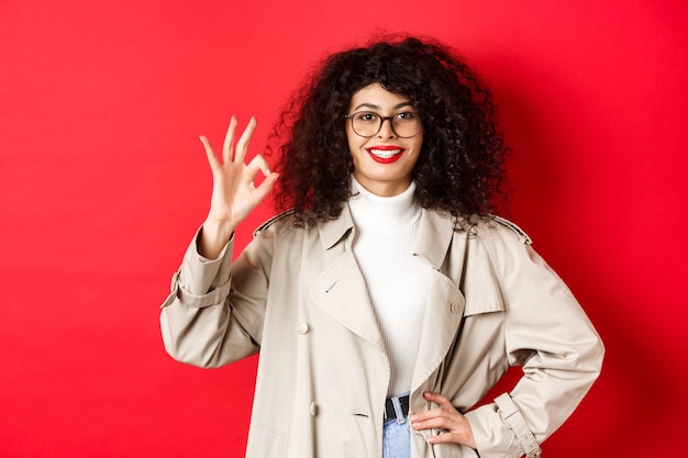 Retrato de mulher confiante na moda de óculos e sobretudo, mostrando o gesto certo para aprovar ou concordar com você, diga sim, de pé na parede vermelha.