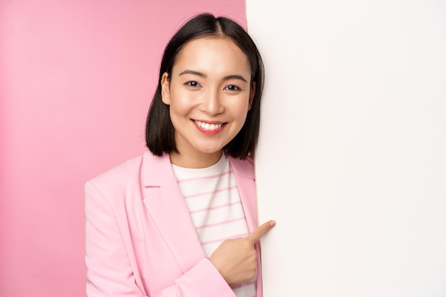 Retrato de mulher corporativa japonesa sorridente feliz apontando o dedo para a placa mostrando smth no espaço vazio em pé de terno sobre fundo rosa