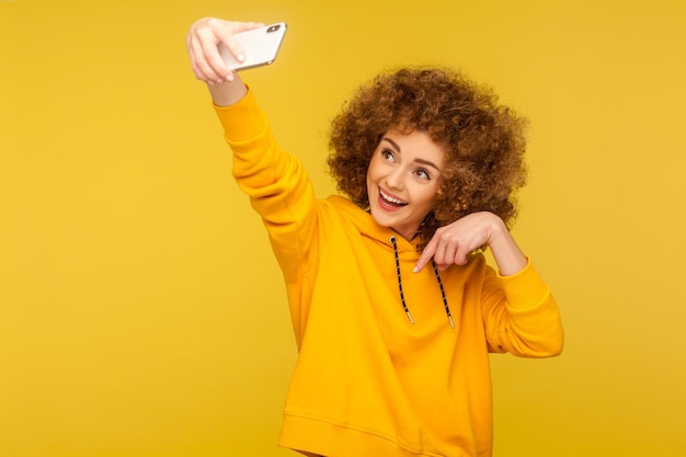 Retrato de mulher de cabelos cacheados na moda alegre com capuz estilo urbano tomando selfie streaming vlog e apontando para baixo pedindo para assinar seu estúdio de rede social isolado em fundo amarelo