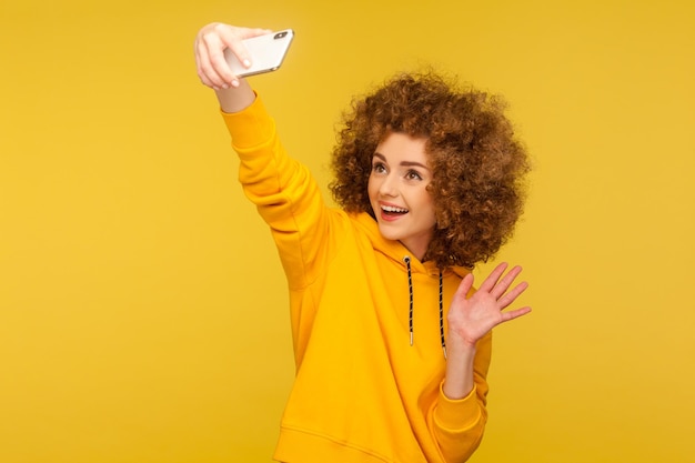 Retrato de mulher de cabelos cacheados na moda feliz com capuz estilo urbano tomando selfie conversando em videochamada e acenando olá gesto vlog estúdio de transmissão tiro isolado em fundo amarelo