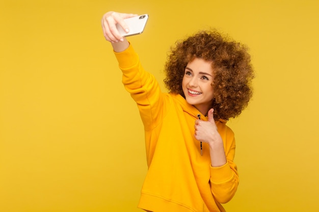 Retrato de mulher de cabelos encaracolados na moda otimista com capuz estilo urbano tomando selfie conversando em videochamada e mostrando os polegares como um estúdio de gestos isolado em fundo amarelo