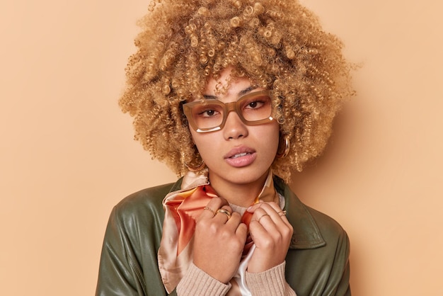 Retrato de mulher de cabelos encaracolados sério usa óculos sas tem visão ruim olha diretamente para a câmera vestida com poses de jaqueta de couro contra fundo marrom. Expressões faciais e estilo de pessoas