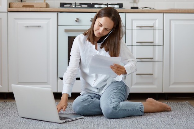 Retrato de mulher de cabelos escuros caucasiana concentrada, vestindo jeans e camisa, sentado contra a cozinha branca definida no chão, usando o computador portátil e falando através do telefone móvel e segurando o documento.