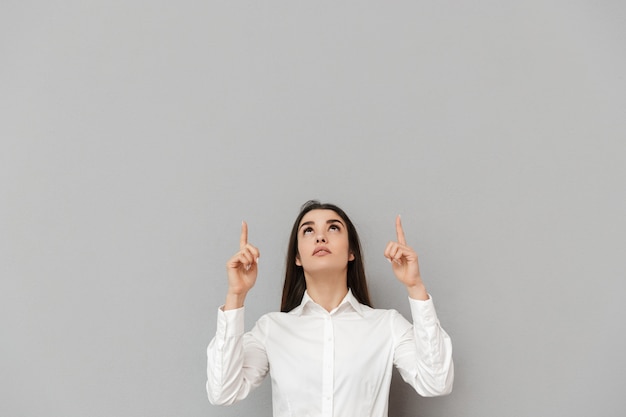 Foto retrato de mulher de escritório inteligente com longos cabelos castanhos em camisa branca, olhando para cima e apontando os dedos para cima na copyspace, isolado sobre a parede cinza
