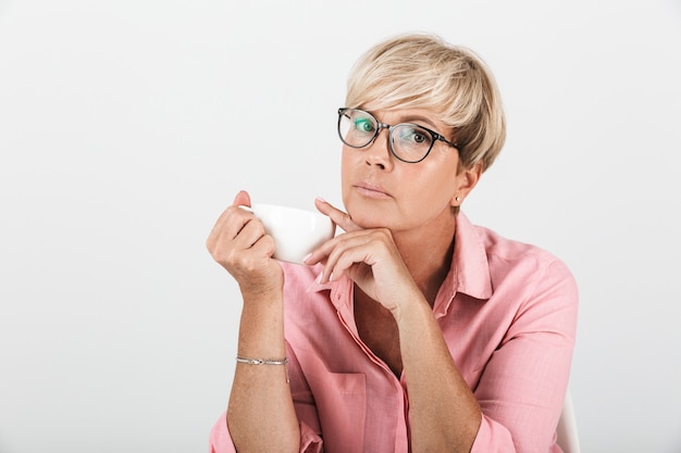 Retrato de mulher de meia-idade concentrada usando óculos, olhando para a câmera e segurando uma xícara de café isolada na parede branca