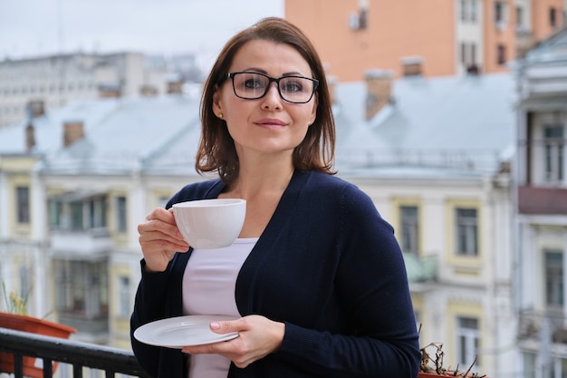 Retrato de mulher de meia-idade em copos, casaco de lã, com uma xícara de chá na varanda aberta na cidade. Arquitetura urbana de fundo, edifícios, mulher sorridente olha para a câmera