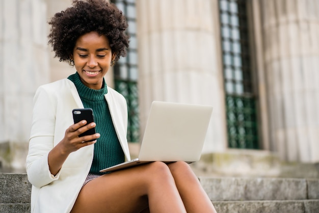 Retrato de mulher de negócios afro usando seu celular e laptop enquanto está sentada na escada ao ar livre