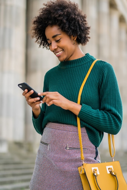 Retrato de mulher de negócios afro usando seu telefone celular em pé ao ar livre na rua. negócios e conceito urbano.