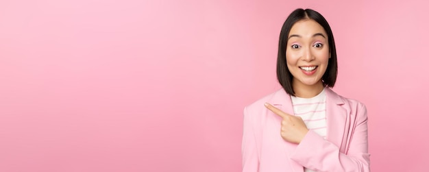 Retrato de mulher de negócios asiática vendedora de terno apontando o dedo para a esquerda mostrando anúncio de banner sorrindo e olhando fundo rosa profissional