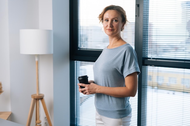 Retrato de mulher de negócios elegante segurando a xícara de café e olhando para a câmera em pé no fundo da grande janela no escritório com luz interior moderna