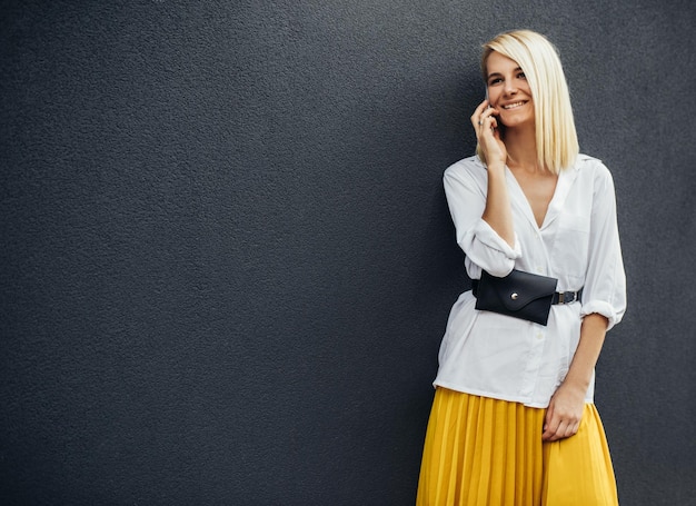 Retrato de mulher de negócios feliz loira sorrindo e de pé contra a parede de edifícios cinza enquanto fala no telefone inteligente Linda mulher caucasiana vestida com camisa branca saia amarela Copiar espaço