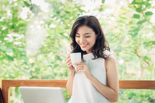 Retrato de mulher de negócios jovem feliz com caneca nas mãos, bebendo café e olhando no laptop.