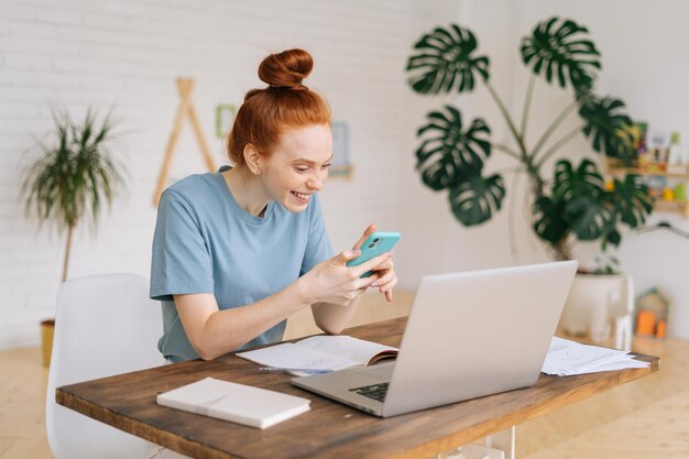 Retrato de mulher de negócios jovem ruiva sorridente feliz usando telefone celular enquanto está sentado na mesa com o laptop na luz aconchegante quarto com interior moderno no escritório em casa