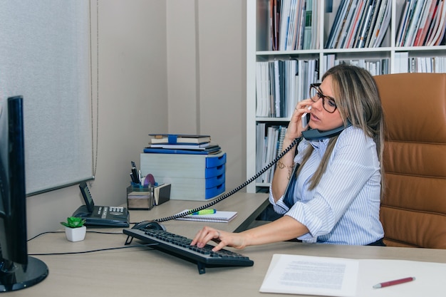 Retrato de mulher de negócios ocupada falando simultaneamente no celular e no telefone fixo