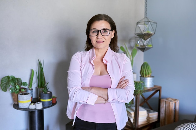 Retrato de mulher de negócios sorridente confiante madura de óculos com os braços cruzados. plantas verdes em vasos, fundo cinza, interior do escritório