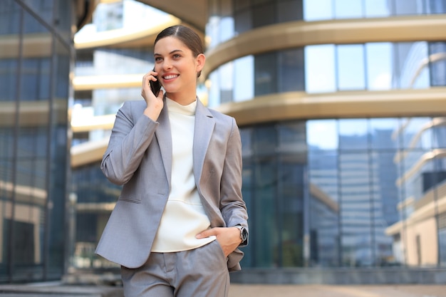 Retrato de mulher de negócios sorridente elegante em roupas da moda, ligando para o celular, perto do escritório.