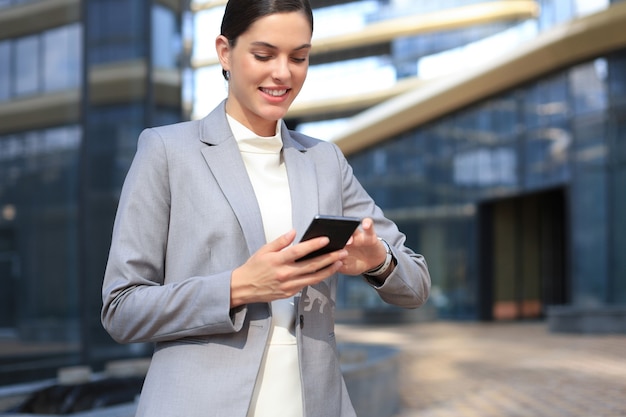 Retrato de mulher de negócios sorridente elegante em roupas da moda, usando telefone celular, perto do escritório.