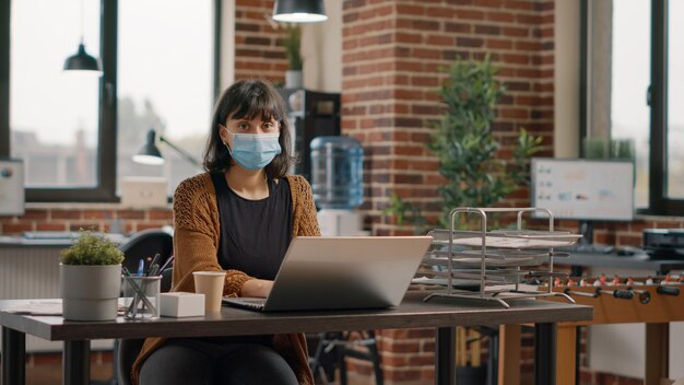 Foto retrato de mulher de negócios, usando máscara facial na mesa e trabalhando com o laptop para projetar o projeto e a estratégia. apresentação do planejamento do empreendedor no computador durante a pandemia de coronavírus.