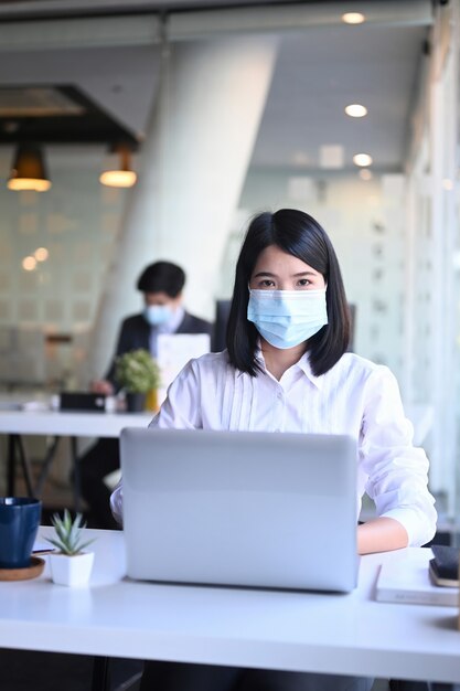 Retrato de mulher de negócios usando máscara protetora, trabalhando no laptop no escritório e seu colega sentado no fundo.