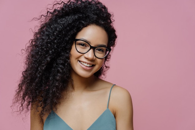 Retrato de mulher de pele escura atraente com cabelo espesso afro vestido casualmente parece através de óculos transparentes tem um sorriso feliz no rosto isolado no fundo rosa Conceito de emoções felizes