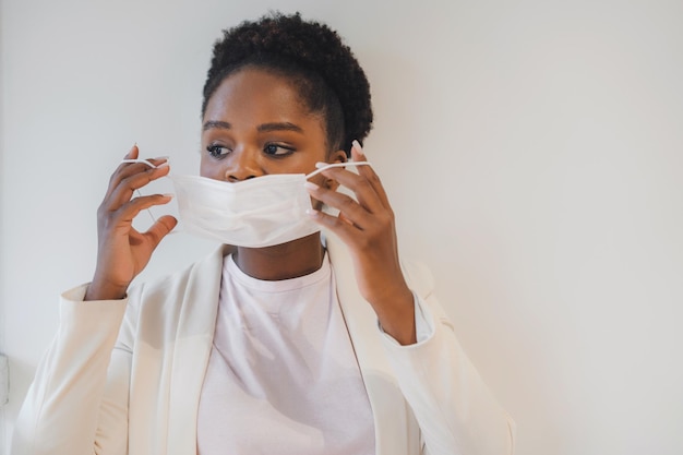 Foto retrato de mulher de pele escura colocando máscara facial médica protetora isolada sobre parede branca novo normal em tempos de coronavírus devido à pandemia de covid19