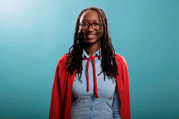 Retrato de mulher de super-herói jovem de aparência positiva vestindo capa vermelha enquanto sorrindo feliz para a câmera sobre fundo azul. Senhora afro-americana orgulhosa e forte vestindo fantasia de herói enquanto está confiante.