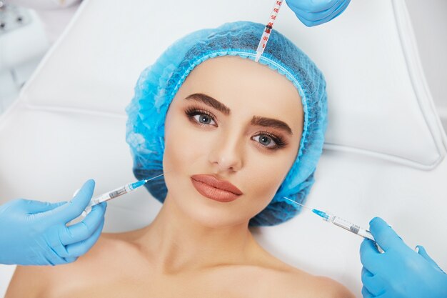 Retrato de mulher deitada no sofá com boné azul na clínica de cosmetologia. Mãos em luvas azuis segurando seringas em volta do rosto do modelo, cosmetologia