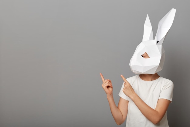 Retrato de mulher desconhecida vestindo camiseta branca e máscara de coelho de papel apontando para o espaço vazio para promoção mostrando a área de anúncio em pé isolada sobre fundo cinza