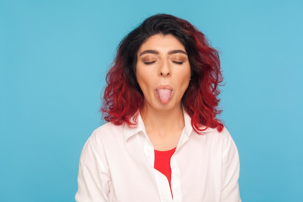 Retrato de mulher desobediente hipster desobediente com cabelo vermelho chique na camisa em pé com os olhos fechados e demonstrando a língua, expressando desobediência. tiro de estúdio interno isolado em fundo azul