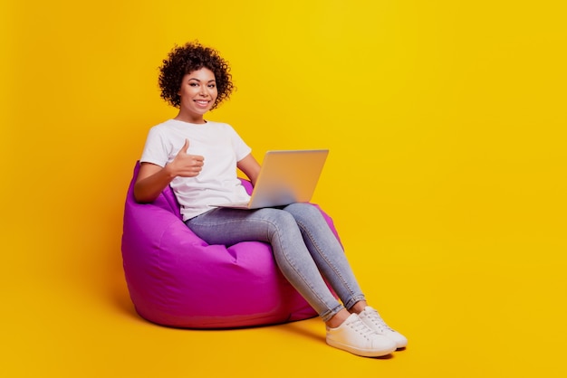 Retrato de mulher digitando em um computador laptop sentar pufe levantar o polegar isolado no fundo amarelo