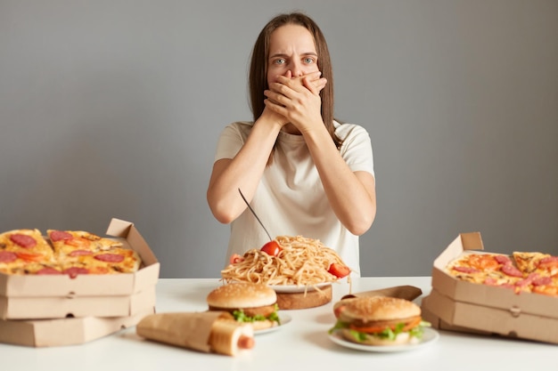 Retrato de mulher doente com cabelo castanho vestindo camiseta branca sentada à mesa isolada sobre fundo cinza sentindo náuseas depois de comer demais junk food