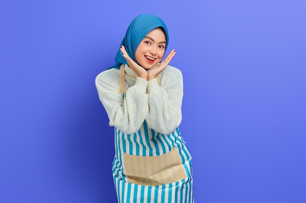 Retrato de mulher dona de casa sorridente em hijab e avental com as duas mãos nas bochechas e olhando para câmera isolada no fundo roxo Conceito de estilo de vida muçulmano dona de casa de pessoas