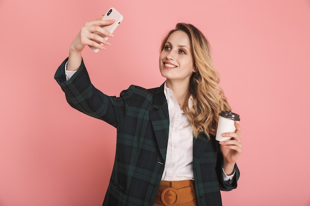 Retrato de mulher elegante de 30 anos em uma jaqueta da moda usando telefone celular e bebendo café para viagem isolado no rosa