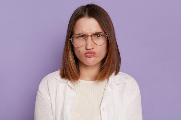 Retrato de mulher engraçada caucasiana, vestindo roupas brancas e óculos, olhando para a câmera com cara engraçada e lábios de beicinho posando isolado sobre fundo roxo