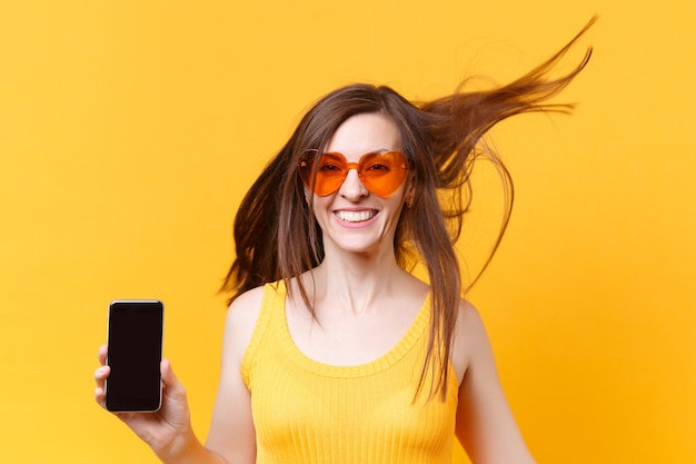 Retrato de mulher engraçada em quadrinhos animado alegre riso em óculos laranja com cabelo esvoaçante segurar o telefone móvel com espaço em branco da cópia de tela vazia isolado no fundo amarelo. Área de publicidade.
