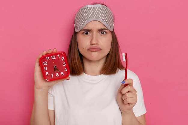 Retrato de mulher engraçada triste chateada com venda nos olhos segurando despertador vermelho e escova de dentes não gosta de limpar os dentes olhando para a câmera com lábios de beicinho tempo para procedimentos matinais