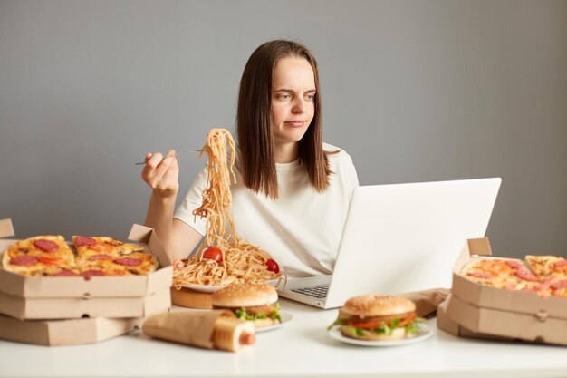 Retrato de mulher entediada insatisfeita assistindo filme no computador portátil comendo macarrão isolado sobre fundo cinza sentado à mesa entre alimentos pouco saudáveis