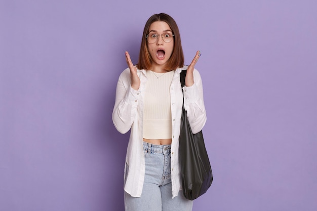 Retrato de mulher excitada chocada com bolsa preta, vestindo camisa branca e jeans posando isolado sobre fundo roxo, olhando para a câmera com a boca aberta e braços erguidos