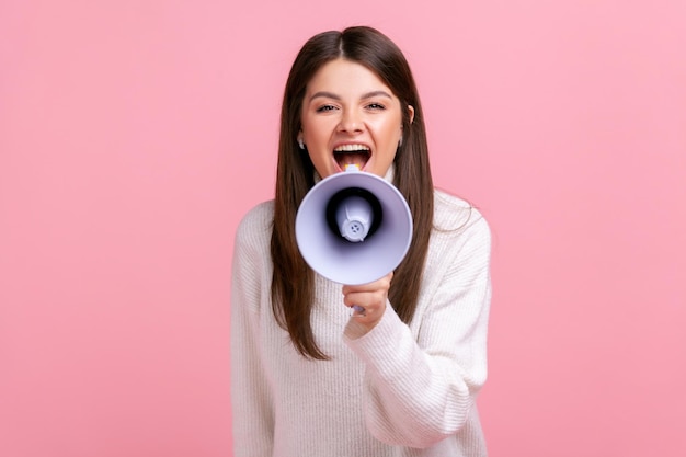 Retrato de mulher excitada gritando no megafone anunciando informações importantes com positivo vestindo suéter branco estilo casual tiro de estúdio interior isolado no fundo rosa