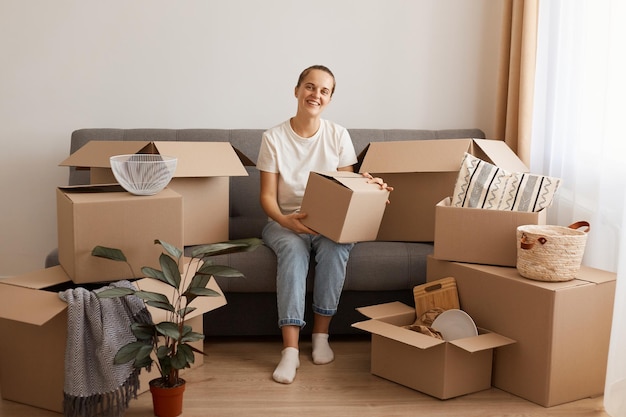 Retrato de mulher feliz atraente vestindo camiseta branca e jeans sentado na tosse em um novo apartamento depois de se mudar, olhando para a câmera com sorriso e expressão feliz, segurando a caixa de papelão.