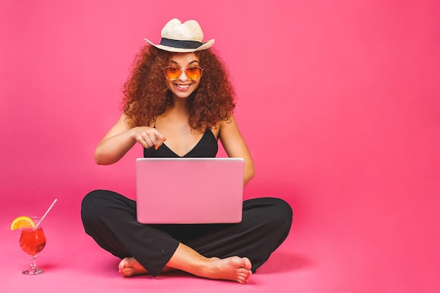 Retrato de mulher feliz casual sentado no chão em posição de lótus e segurando laptop e coquetel isolados