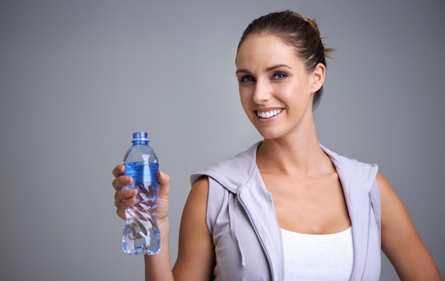 Retrato de mulher feliz e garrafa de água em aptidão para hidratação em um fundo de estúdio cinza Rosto de pessoa feminina ou atleta com sede com sorriso e bebida mineral para sustentabilidade natural em maquete